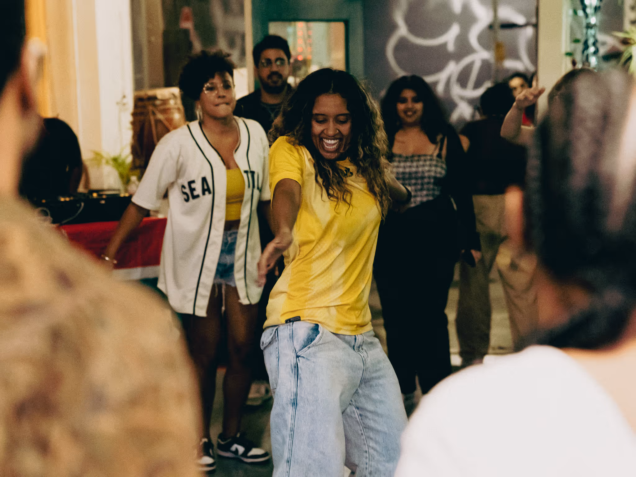 Una persona vestida con una camiseta amarilla y unos vaqueros azul claro baila en el centro de una pequeña multitud, sonriendo e inclinándose hacia adelante con un brazo extendido como si estuviera a mitad de un paso. Varias personas se paran detrás de ellas, observándolas y moviéndose, vestidas de manera informal con camisetas, pantalones cortos y zapatillas deportivas. La escena tiene lugar en interiores con una iluminación cálida, plantas y un mural o una obra de arte visible en la pared. La multitud forma un círculo suelto alrededor del bailarín, creando una atmósfera social y enérgica centrada en el movimiento y la música.