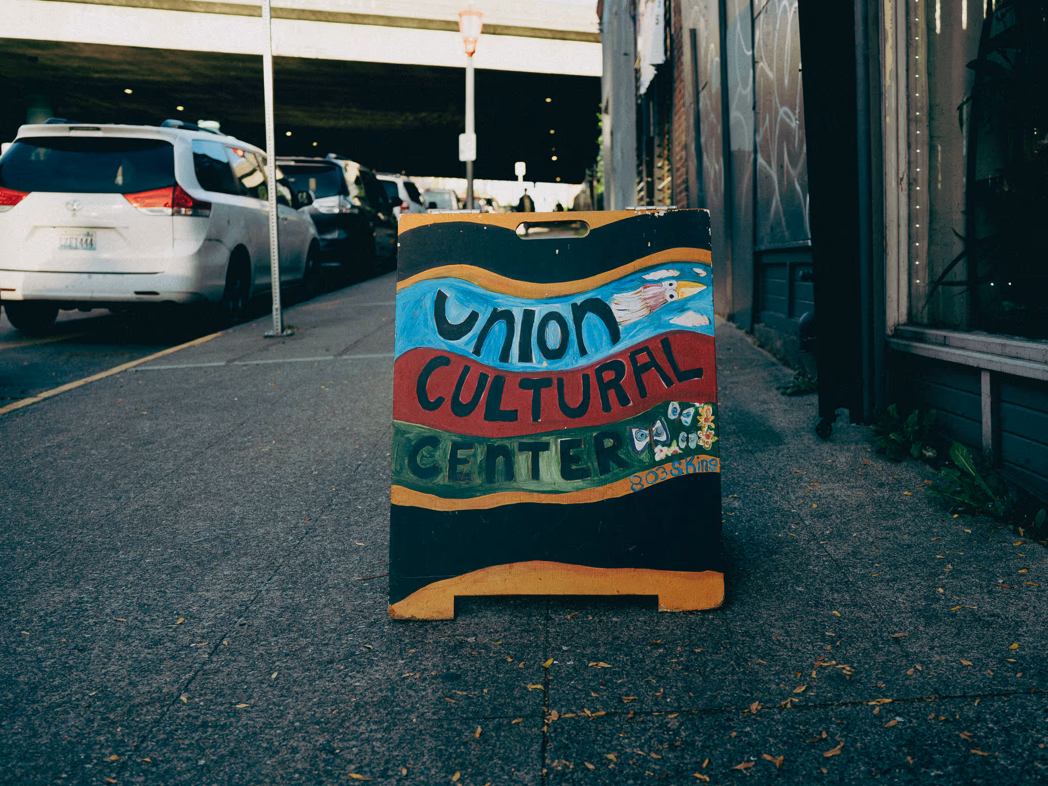 Un letrero pintado a mano se encuentra en una acera de concreto a lo largo de una calle de la ciudad. El letrero dice «Union Cultural Center» en letras grandes y coloridas, con franjas azules, rojas y verdes fluidas y pequeños detalles ilustrados. Los automóviles estacionados se alinean en la calle a la izquierda y, al fondo, un paso elevado o una carretera elevada se extiende por encima. A la derecha, una pared de ladrillo con grafitis y una puerta oscura enmarca la acera. Las hojas caídas están esparcidas por el suelo y el letrero se coloca como señal de bienvenida fuera del edificio.