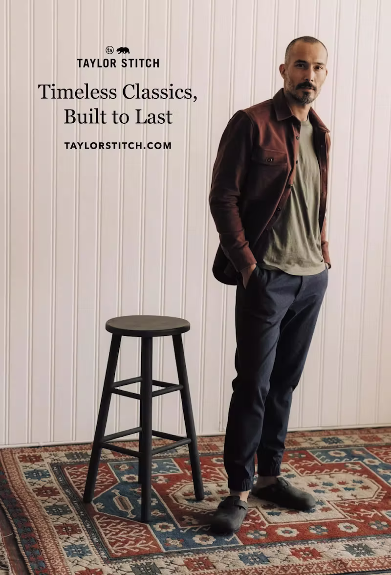 A minimalist-style postcard showing a man in a rust jacket, olive tee, and navy pants standing confidently beside a black wooden stool on a patterned rug. The left side features the Taylor Stitch logo and tagline: “Timeless Classics, Built to Last” with their website: taylorstitch.com.