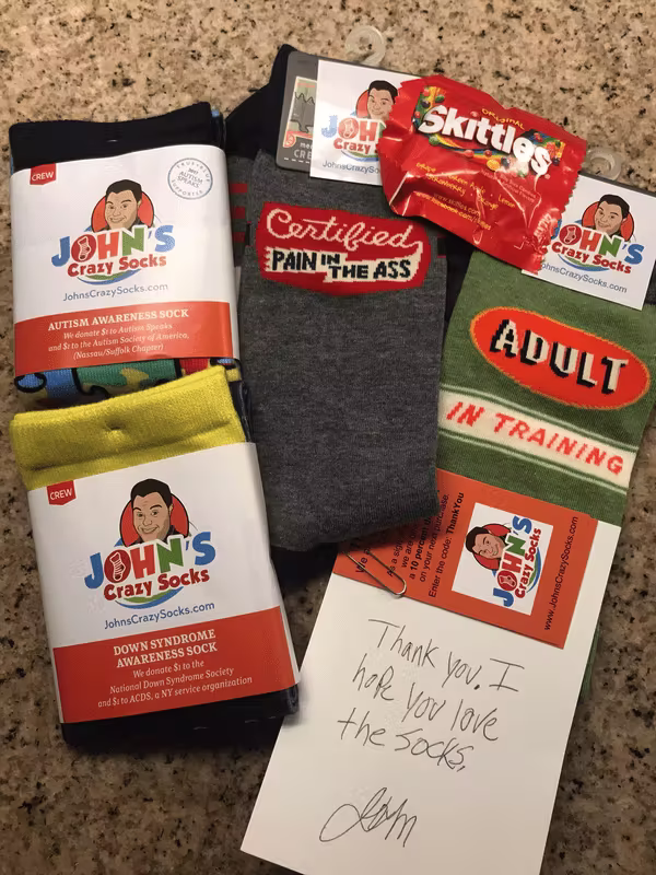 Assortment of colorful crew socks and packaging from John’s Crazy Socks spread on a granite countertop. Visible pairs include: a blue pair labeled “AUTISM AWARENESS SOCK,” a yellow/blue pair labeled “DOWN SYNDROME AWARENESS SOCK” (with a note on the label about donations to the National Down Syndrome Society), a gray pair with a bold woven phrase “Certified PAIN IN THE ASS,” and a green/orange pair reading “ADULT IN TRAINING.” Brand labels show a cartoon portrait logo and “John’s Crazy Socks.” A mini red pack of Skittles candy lies on top. A small white card with quick handwriting says: “Thank you. I hope you love the socks. —John” with a cursive signature.