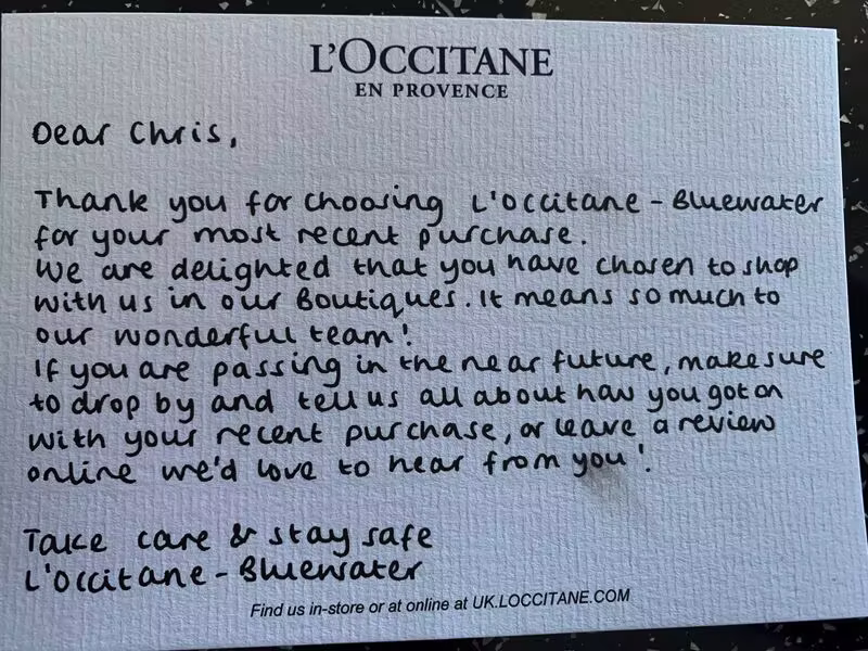 Textured cream card with the navy “L’OCCITANE EN PROVENCE” logo centered at the top. Neat black-ink handwriting reads: “Dear Chris, Thank you for choosing L’Occitane – Bluewater for your most recent purchase. We are delighted that you have chosen to shop with us in our Boutiques. It means so much to our wonderful team! If you are passing in the near future, make sure to drop by and tell us all about how you got on with your recent purchase, or leave a review online we’d love to hear from you! Take care & stay safe L’Occitane – Bluewater.” Small printed footer at the bottom: “Find us in-store or online at UK.LOCCITANE.com.” The card sits on a dark, slightly speckled surface.