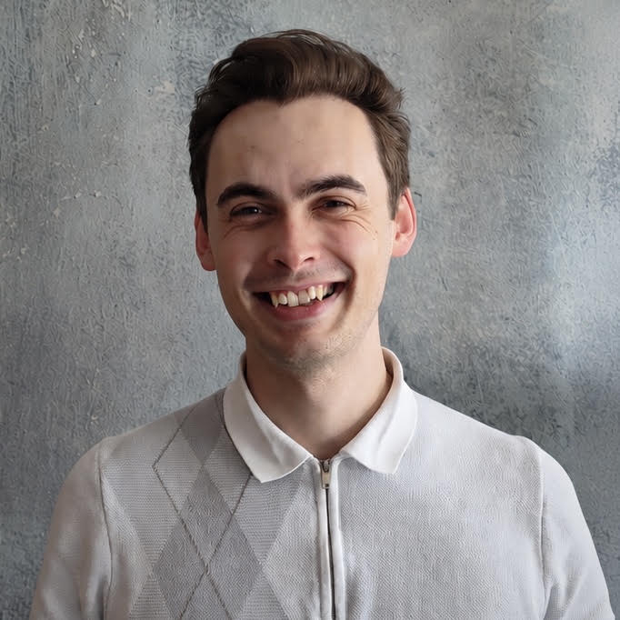 Smiling young man with dark hair wearing a light-colored sweater against a textured gray wall.