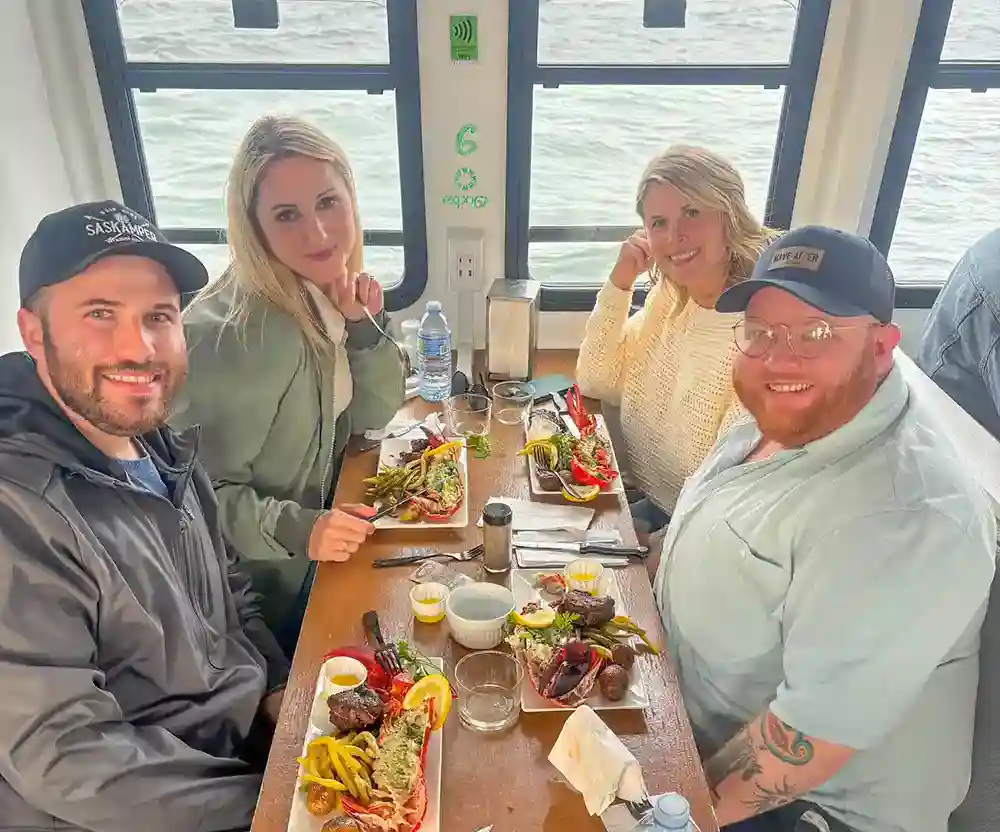 Four guests enjoy the Surf n Turf Dinner Cruise in Charlottetown, PEI. They are looking at the camera, smiling, meals in front of them, the Charlottetown Harbour is seen in the background.