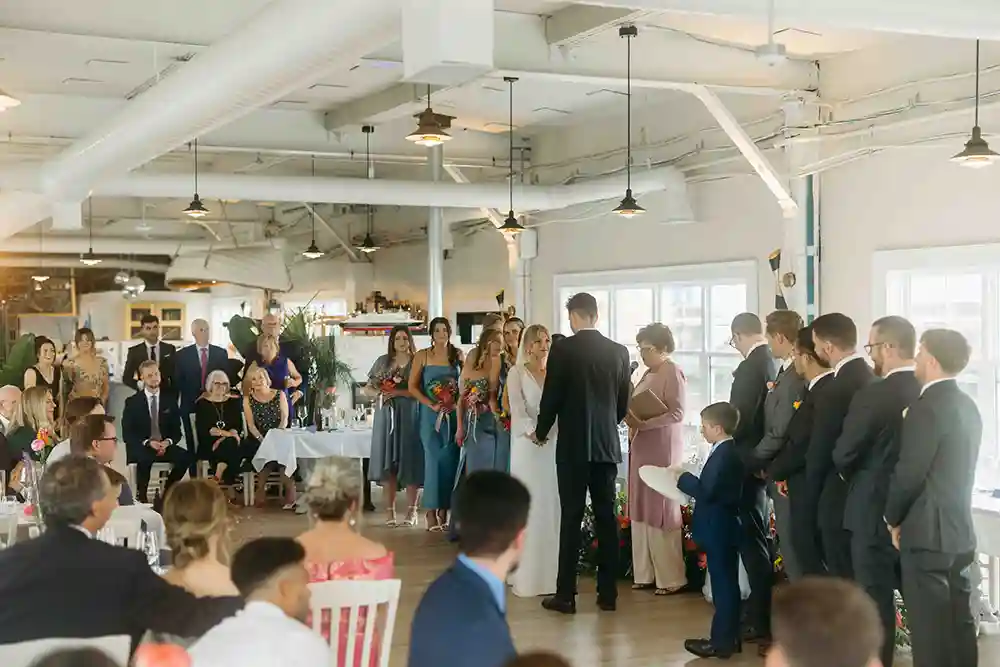 A bride and groom share vows during their wedding day at Cable Wharf Restaurant. A crowd of guests look on.