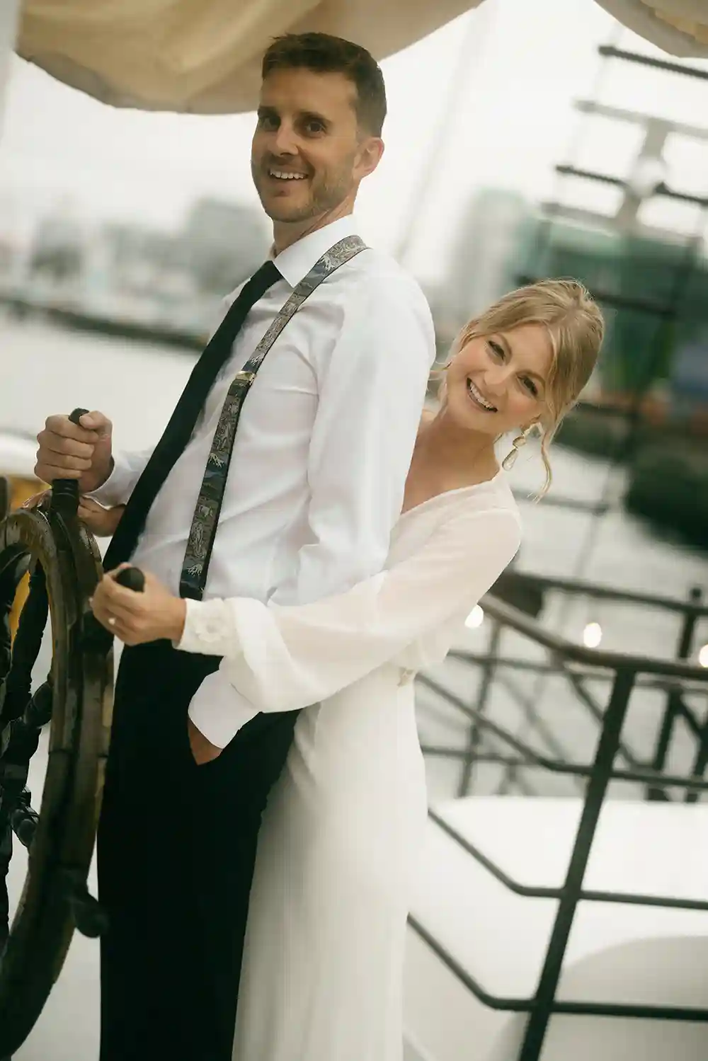 Newlyweds onboard Tall Ship Silva. The husband holds the ships steering wheel while the bride is hugging him. They're smiling.