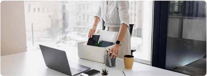 Person packing up office supplies including laptop, plant, and notebook into a white box on a desk.
