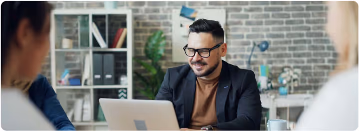 Man wearing glasses and a blazer working on a laptop at a desk with blurred colleagues in the foreground.