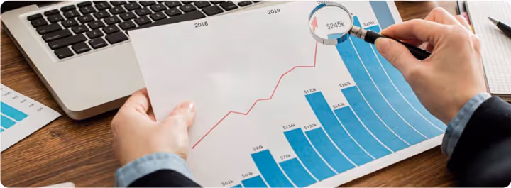 Person holding a financial chart with blue bar graphs and a red line graph while examining a $245k value through a magnifying glass with a laptop keyboard in the background.