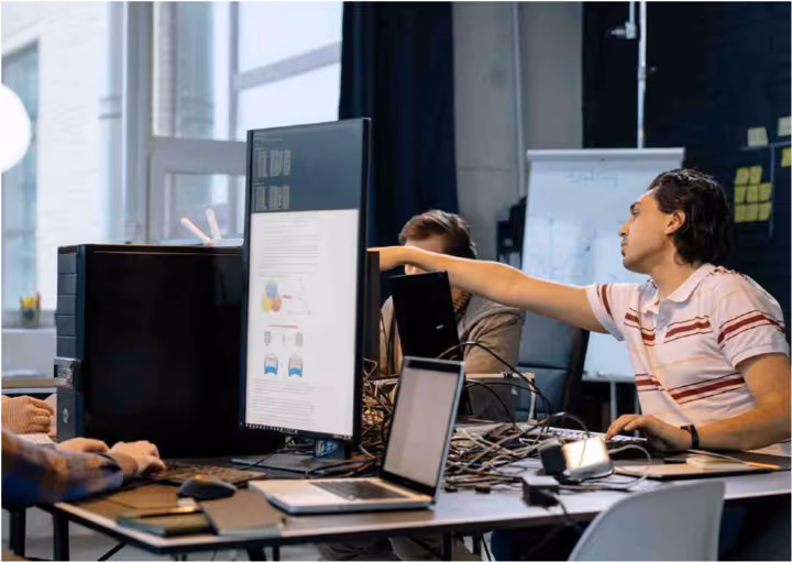 Two men working together at a desk with laptops and a vertical monitor displaying charts and code.