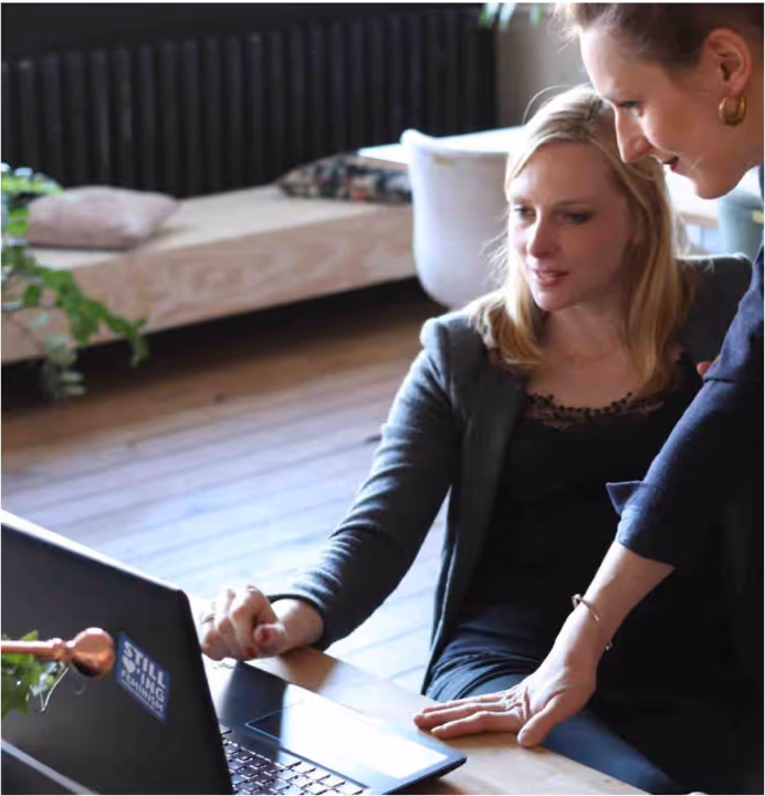 Two women working together at a laptop in a cozy, modern room with wooden flooring and a bench in the background.