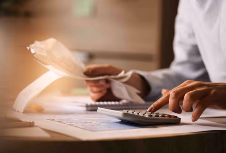 Person using a calculator and holding receipts while managing finances at a desk.