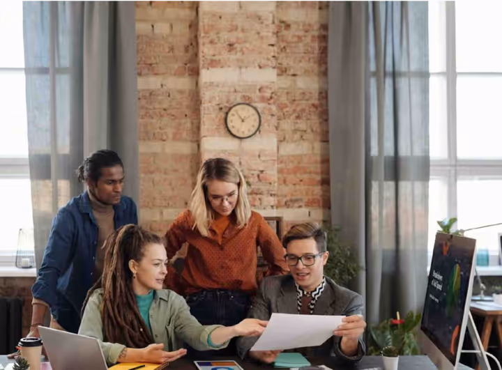 Four young coworkers in a modern office reviewing a document together at a table with a laptop and desktop computer.