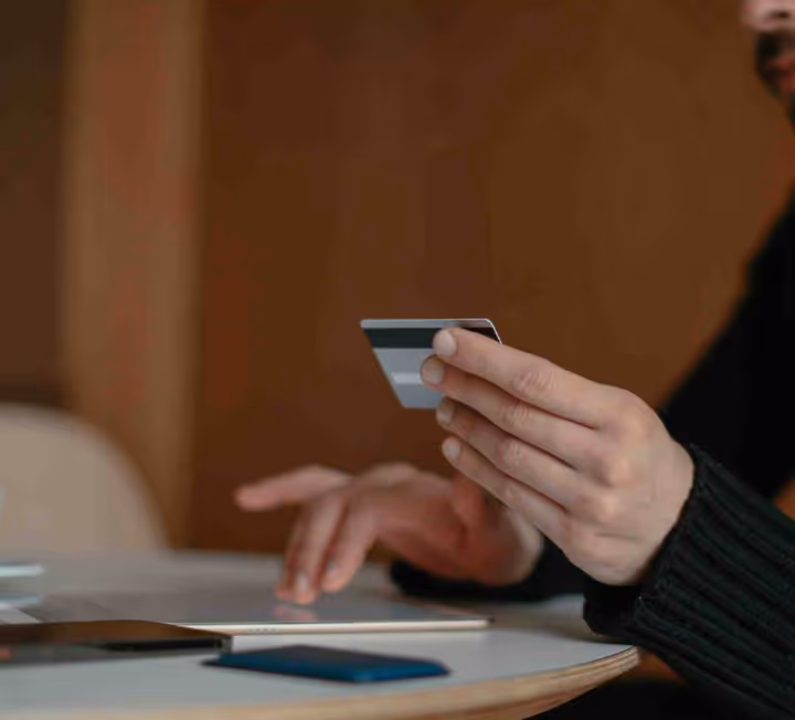 Person holding a credit card and using a tablet on a round table with two smartphones nearby.