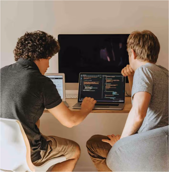 Two young men collaborating on coding at laptops in a casual work setting.