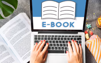 Hands typing on a laptop keyboard with an e-book icon displayed on the screen and an open book nearby.