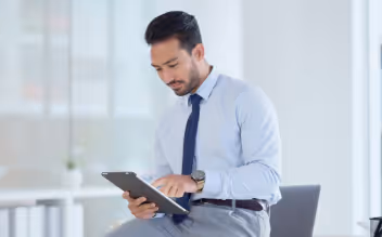 Man in light blue shirt and navy tie sitting and using a tablet in a bright office setting.