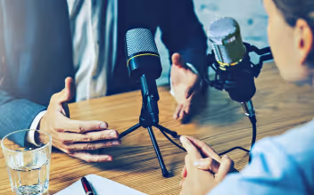Two people sitting at a table having a podcast conversation with microphones and a glass of water.