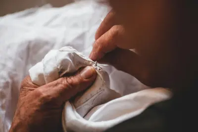 Mãos idosas costurando cuidadosamente um tecido branco com agulha e linha