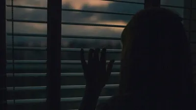 Pessoa observando através de persiana com ambiente escuro ao fundo, remetendo à sensação de medo ou introspecção