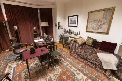 Visão ampla do consultório de Freud com o divã e mesa de trabalho, repleto de esculturas e tapetes orientais.
