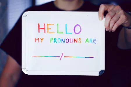 Pessoa segura uma placa com a frase 'Hello, my pronouns are' escrita em letras coloridas, representando a diversidade de identidades de gênero