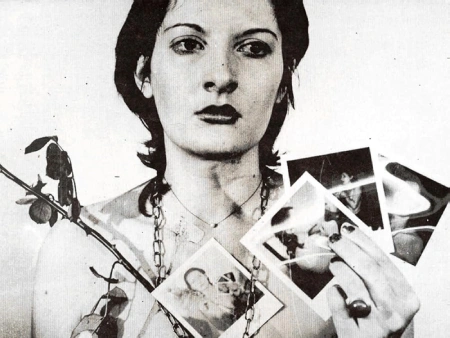 Registro de performance de Marina Abramović segurando fotografias, com elementos naturais e olhar direto para a câmera