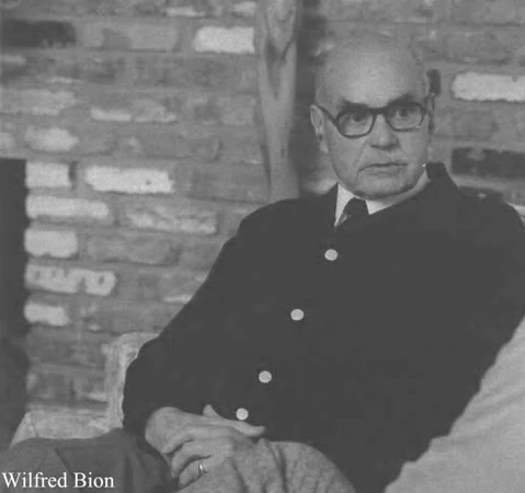 Wilfred Bion sentado em retrato em preto e branco, com fundo de parede de tijolos