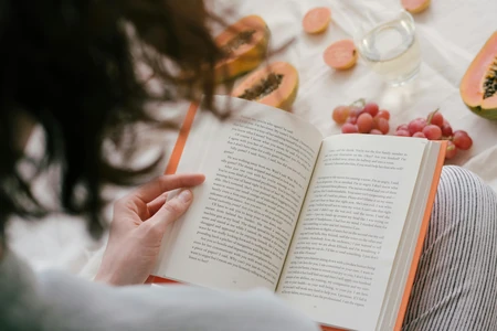 Pessoa lendo um livro ao ar livre com outros objetos sobre uma mesa.