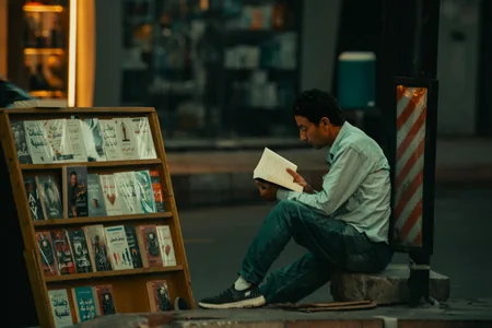 Homem sentado na calçada lendo um livro diante de uma banca