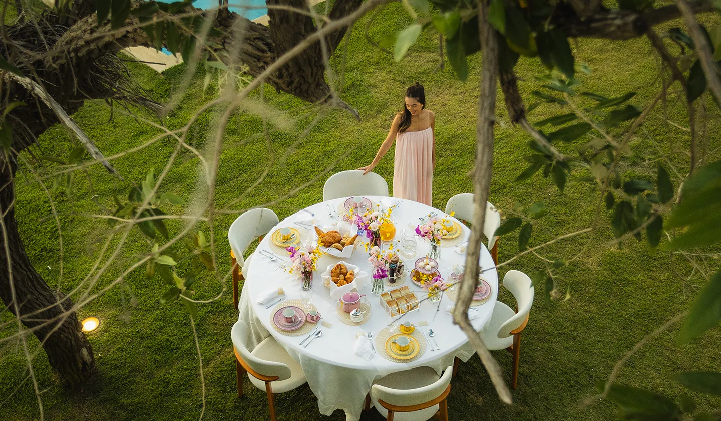Woman in pink dress standing by a round outdoor table set with pastries, flowers, and tea cups.
