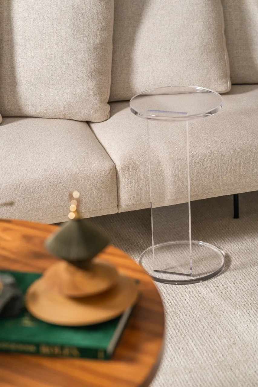 Clear acrylic side table beside a beige textured sofa with cushions on a light carpet.