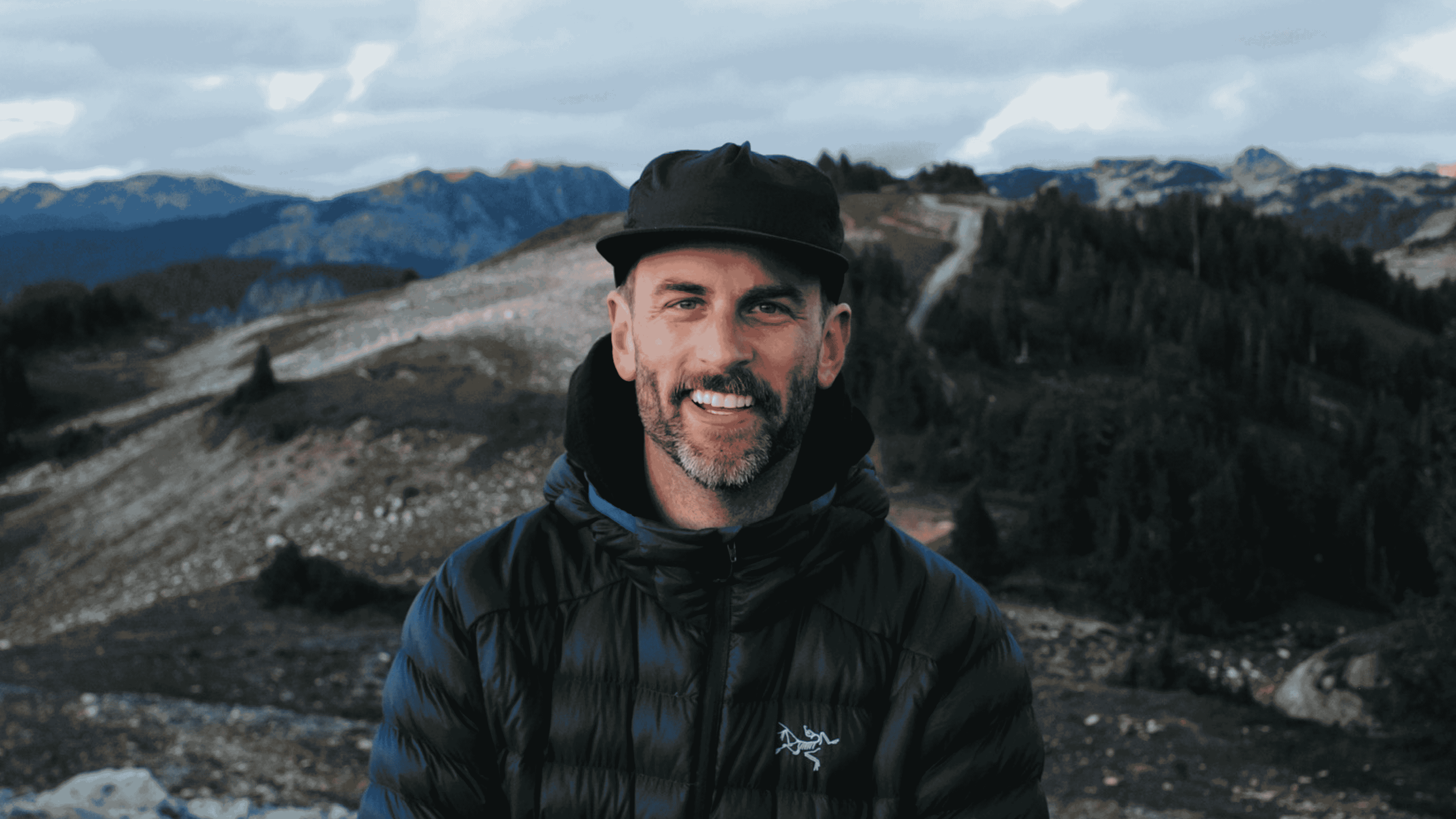 Smiling man with a beard wearing a black cap and puffer jacket outdoors with a mountainous landscape in the background.
