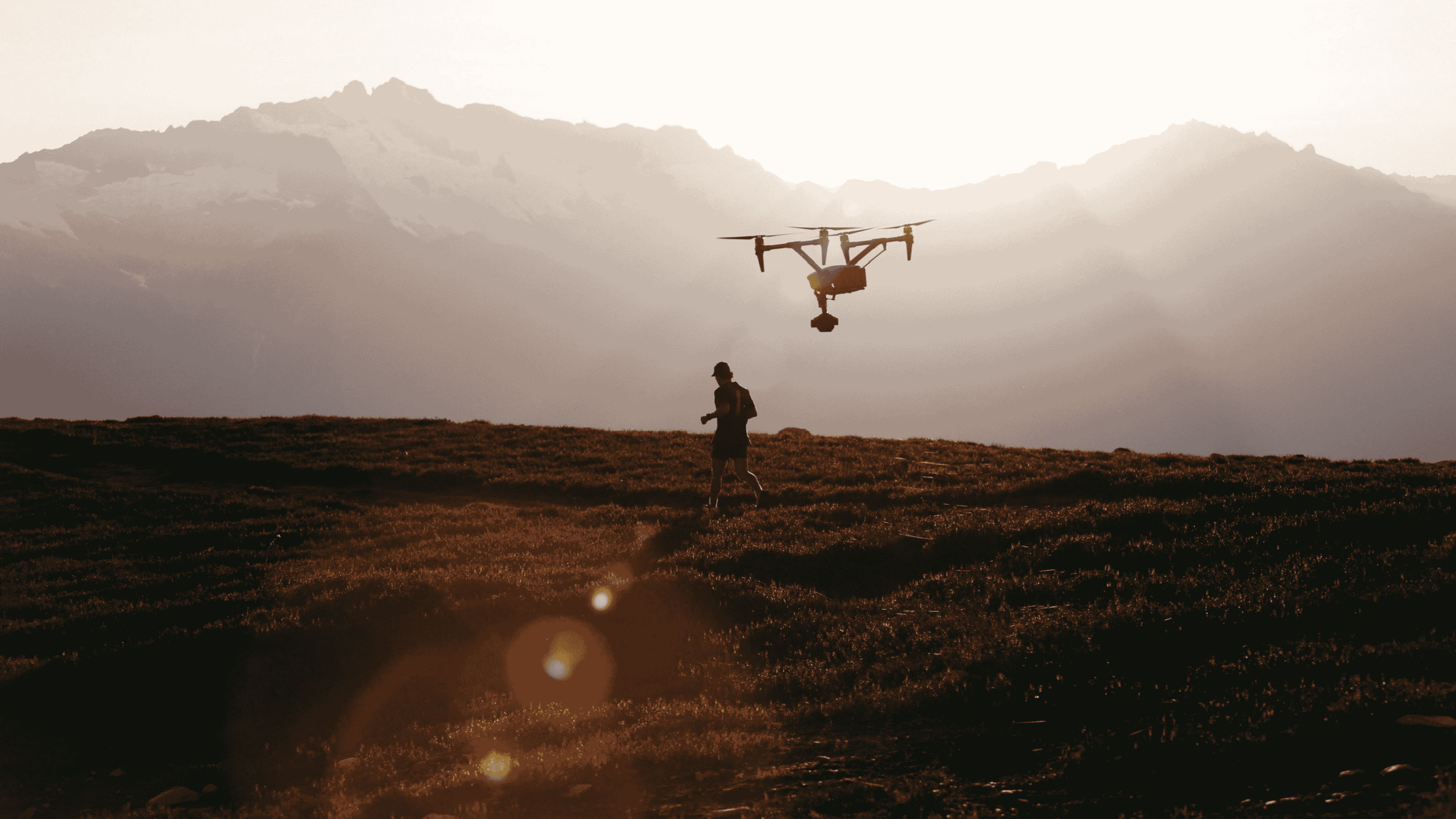 Person walking on grassy terrain with a drone flying nearby against a backdrop of hazy mountains at sunset.
