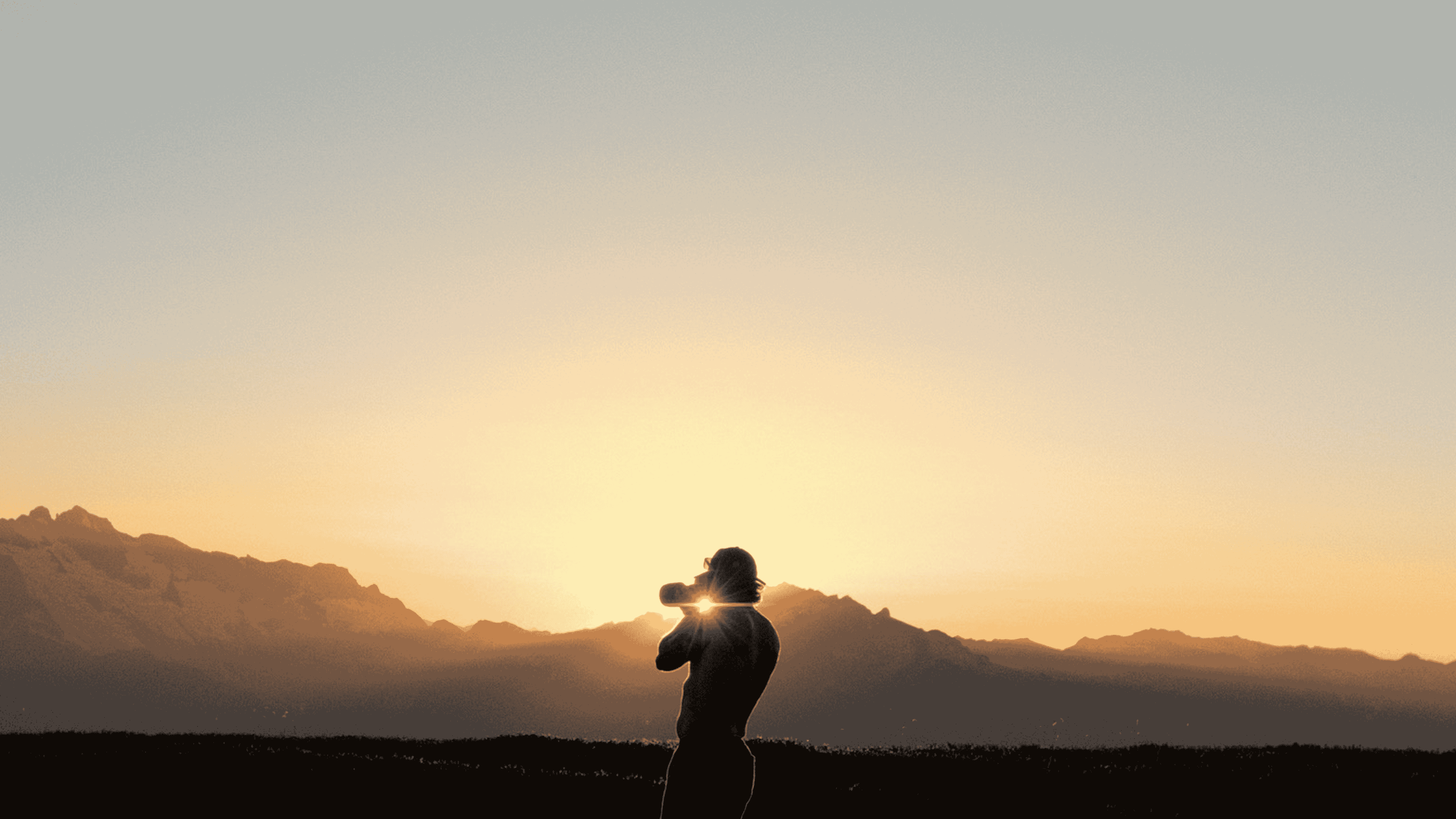 Silhouette of a person drinking from a water bottle with mountains and a sunset in the background.