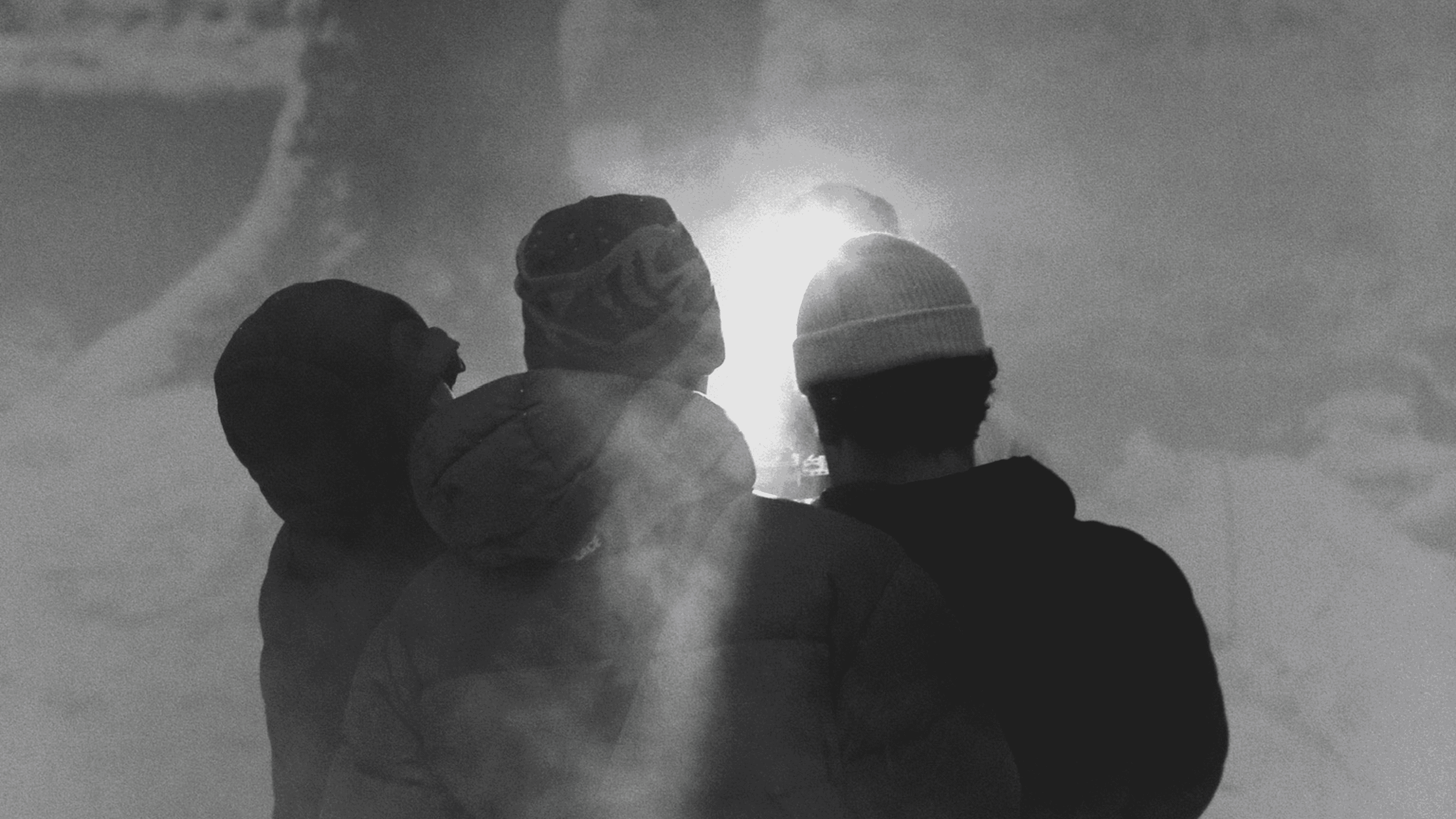 Three people wearing winter jackets and hats standing with their backs to the camera, facing a bright light surrounded by mist or smoke.