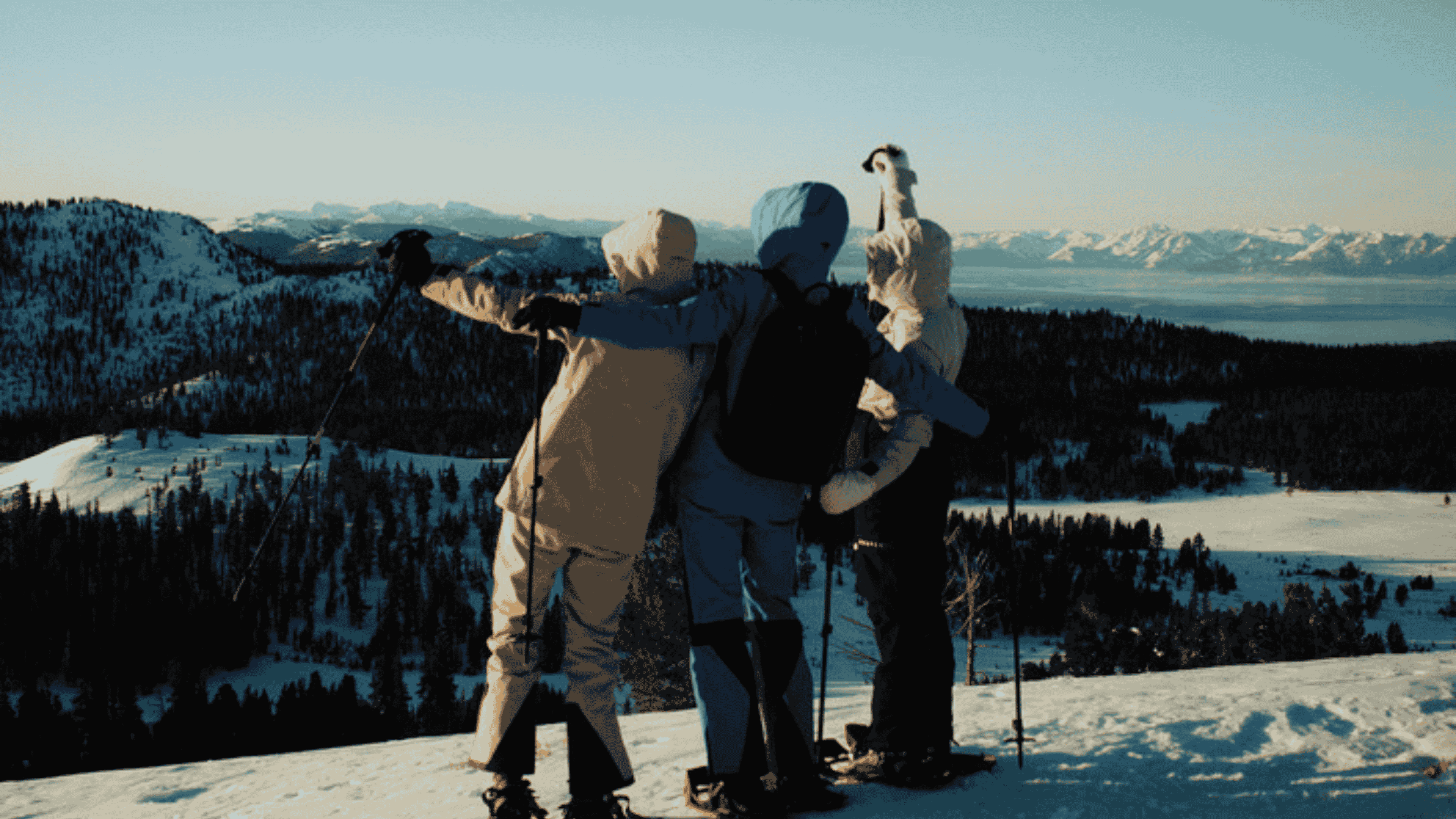 Three skiers standing side by side with arms around each other, overlooking snow-covered mountains at sunset.