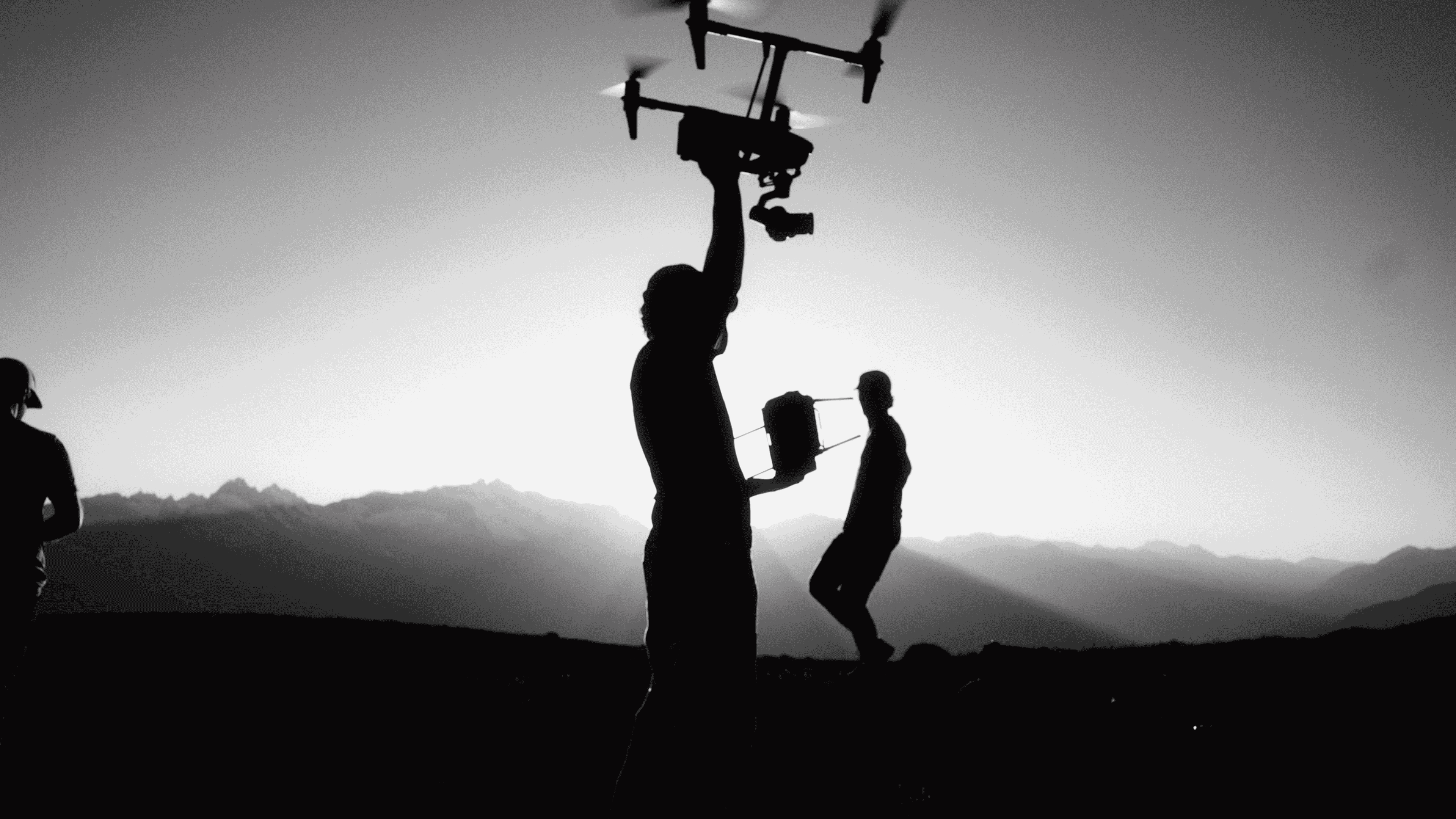 Silhouettes of three people in front of mountains during sunset, one holding a drone above his head and another holding its controller.