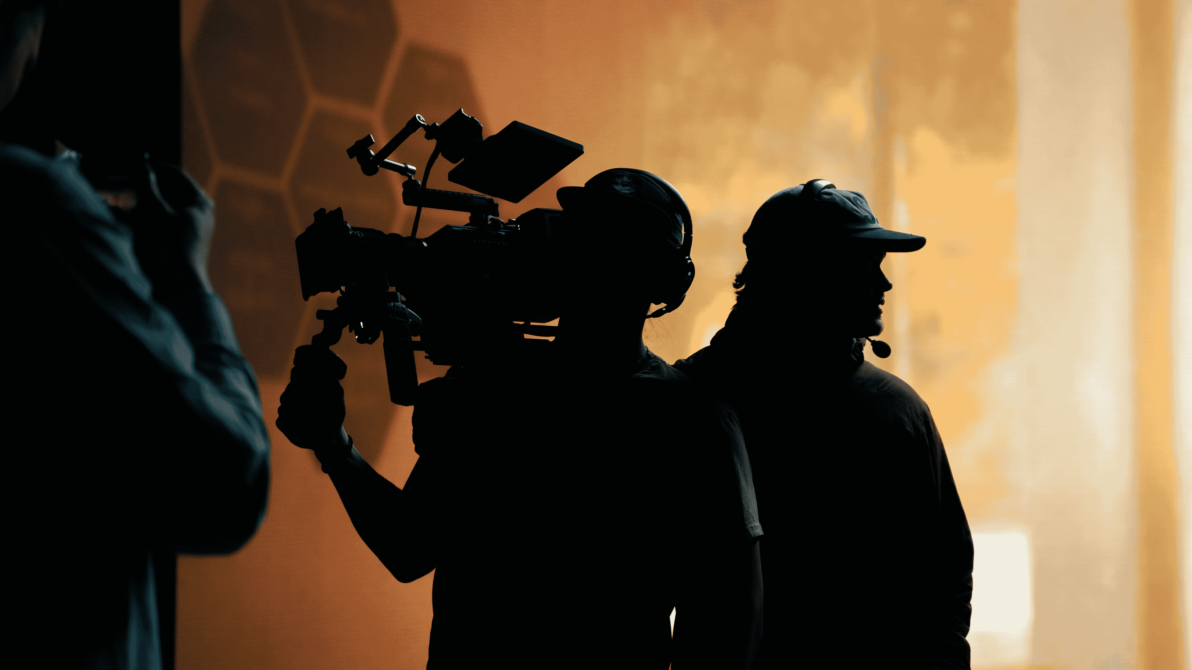 Silhouetted camera operator filming with a video camera and another person wearing a headset in front of an orange-yellow background.