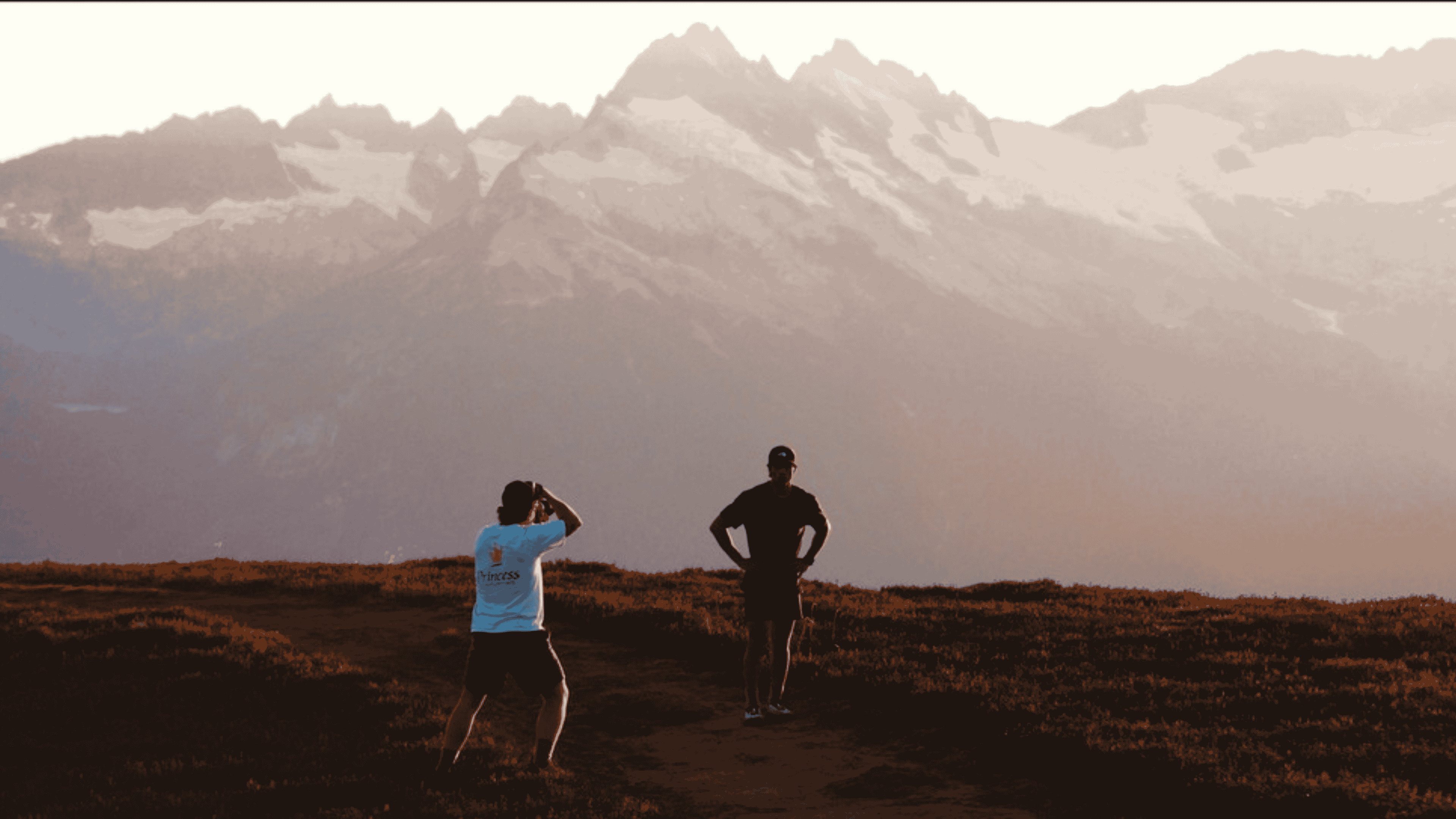 Two people on a dirt path in a mountainous area at sunset, one taking a photo of the other.