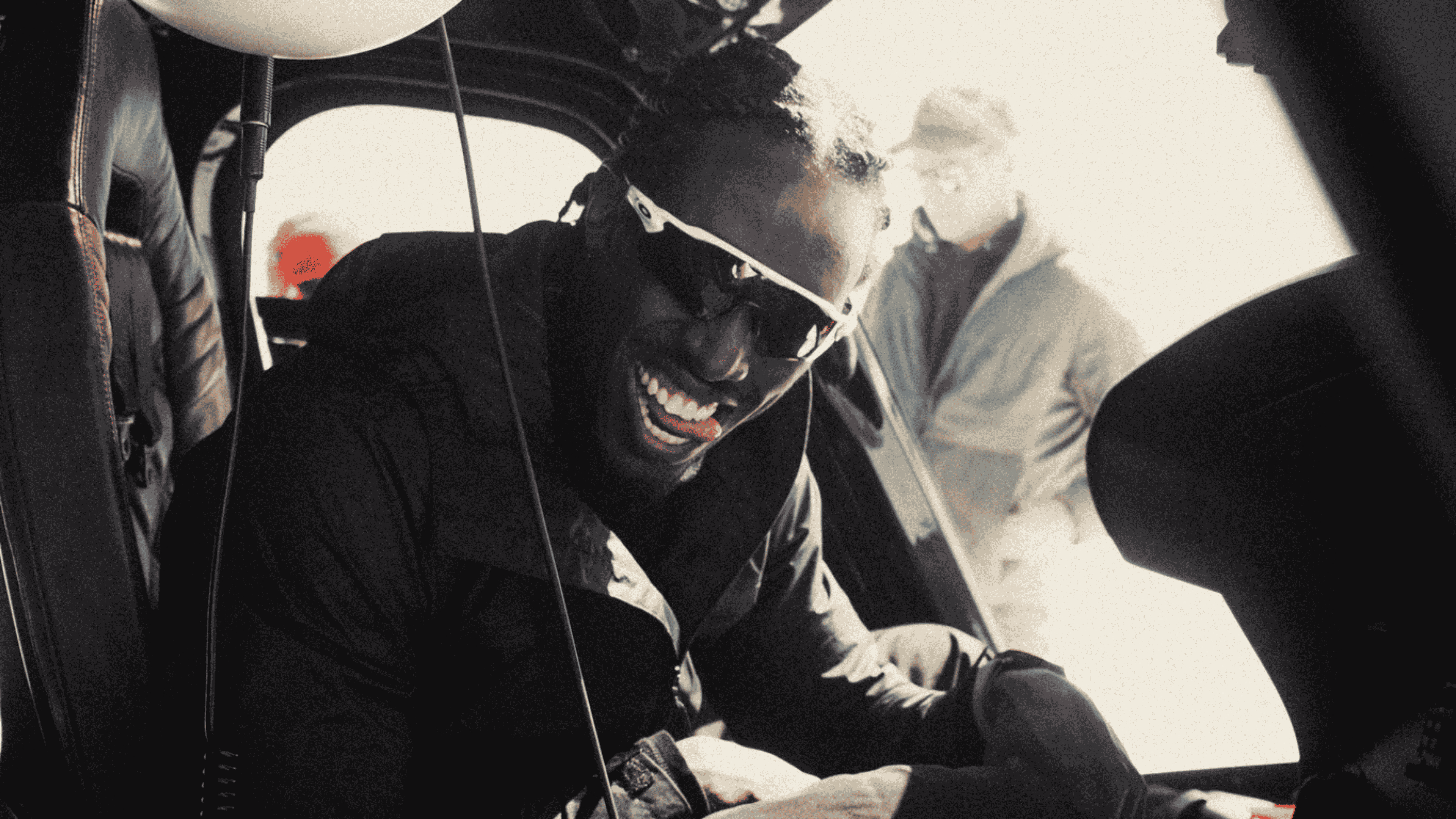 Man wearing sunglasses and a dark jacket smiling inside a vehicle with another person blurred in the background.