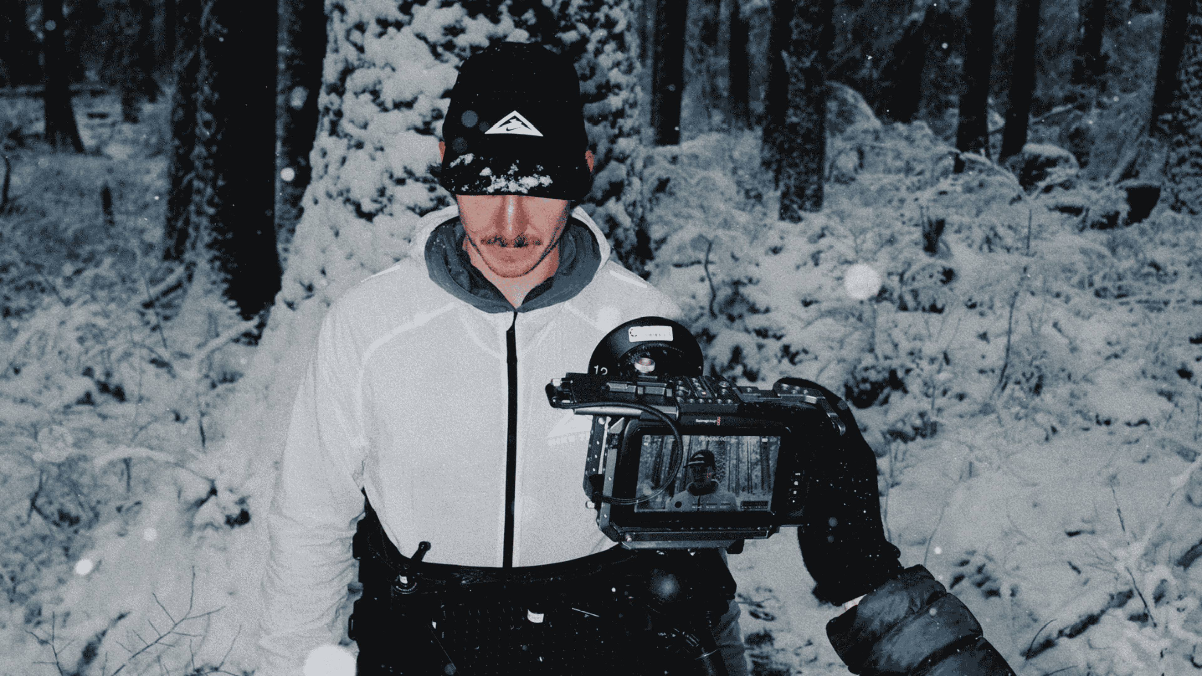 Man wearing a white jacket and black cap standing in a snowy forest while being filmed by a camera held by a person in a black glove.