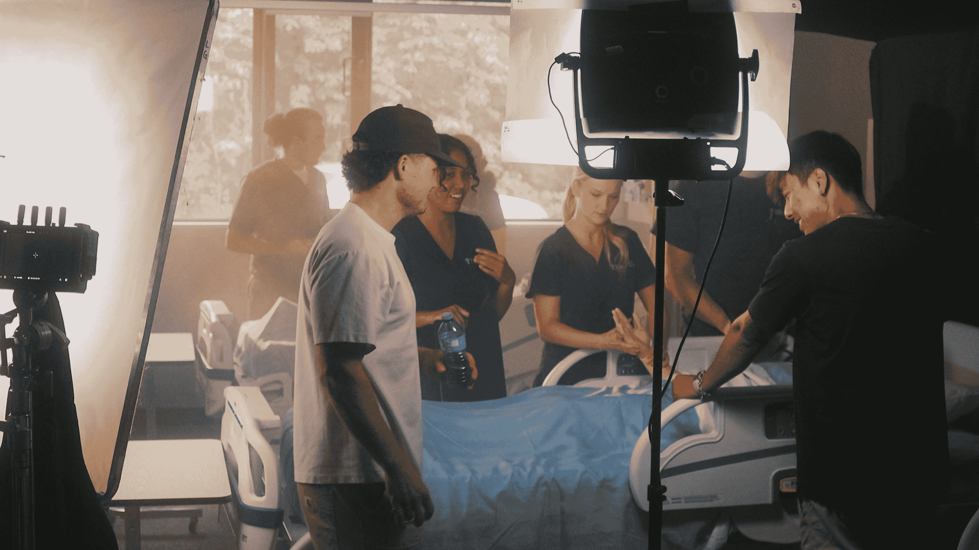Group of people around a hospital bed with filming equipment and lighting setup.