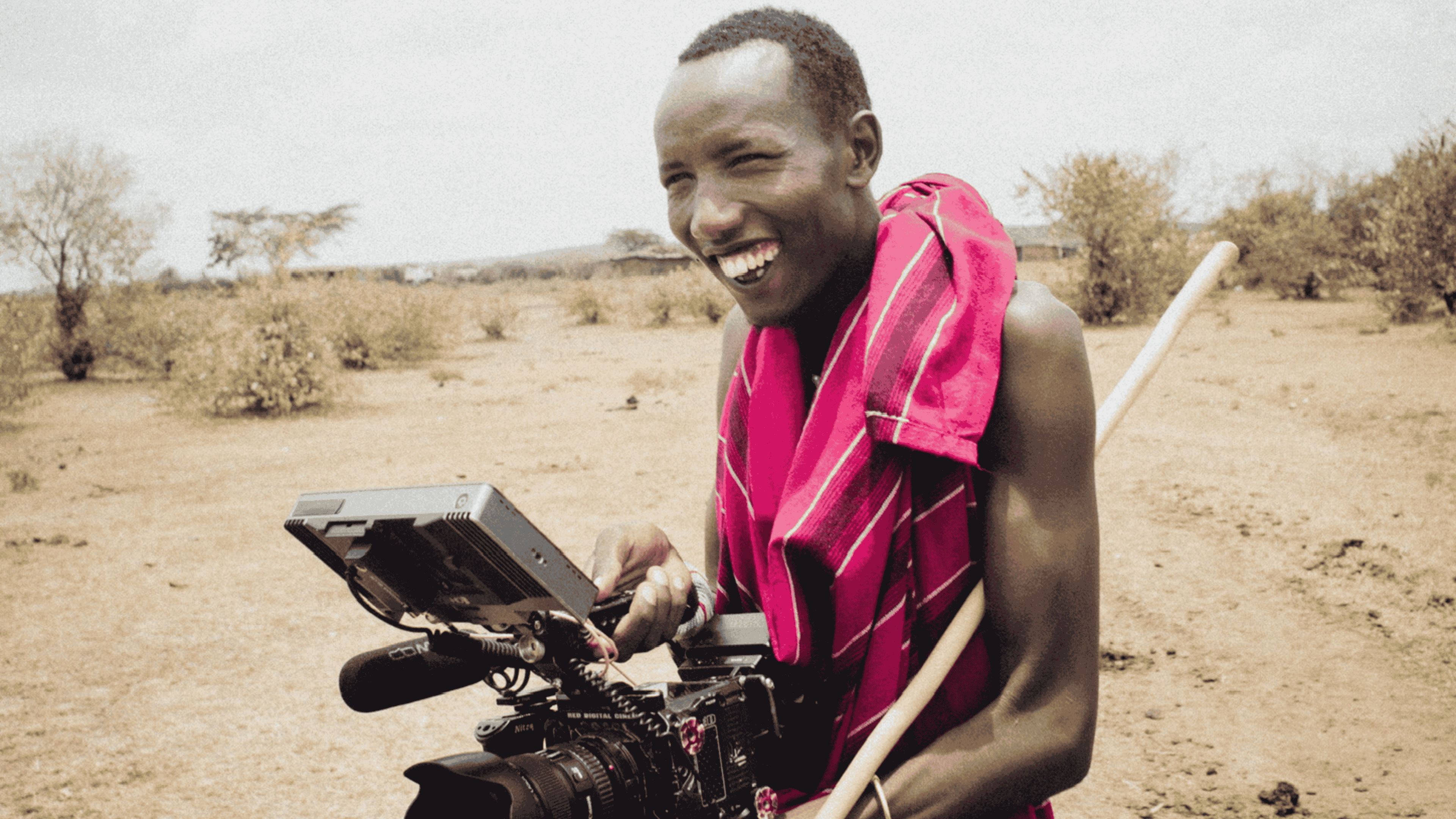 Smiling person wearing a red patterned cloth holding a professional video camera in a dry, bushy landscape.