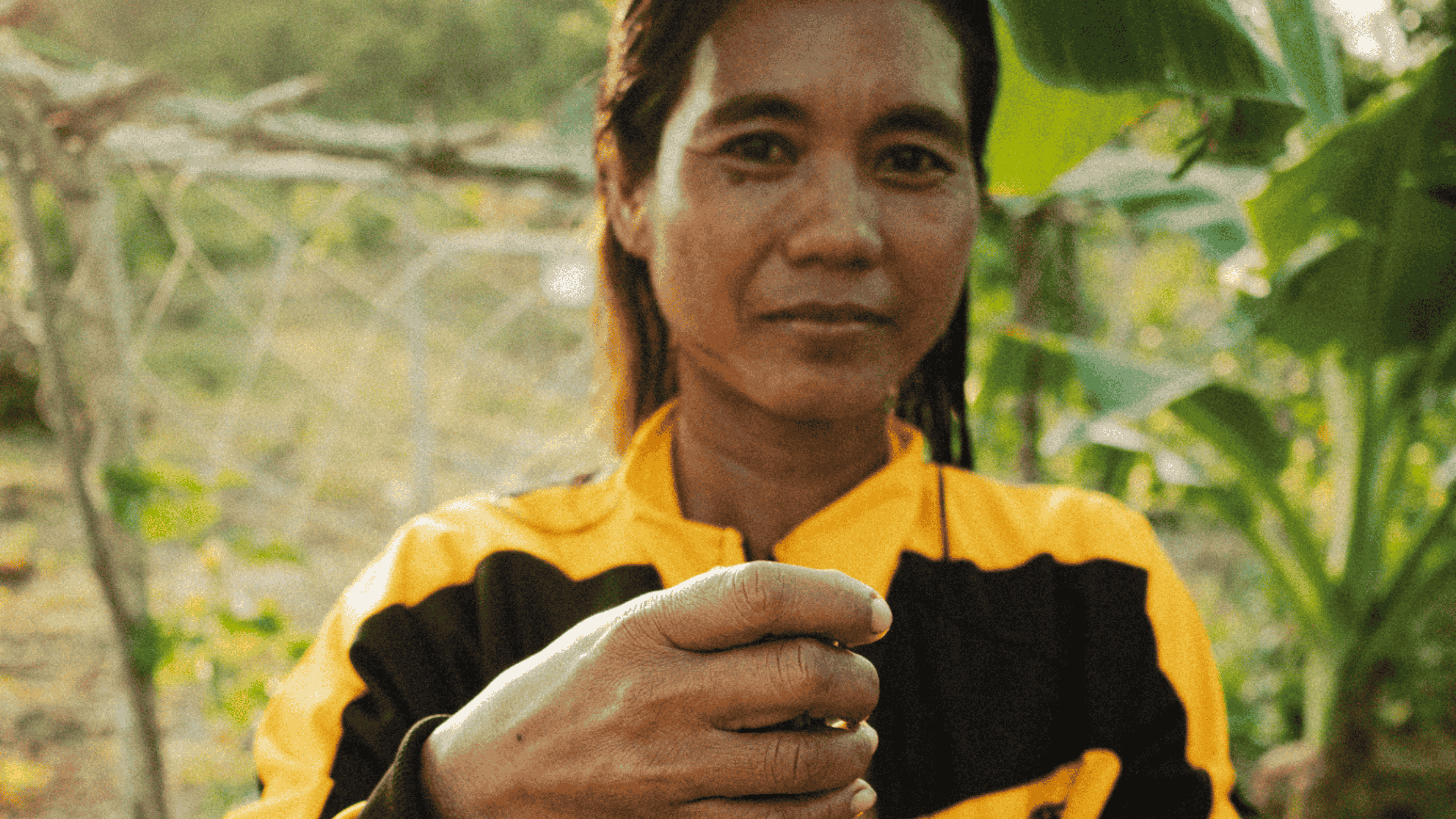 Person wearing a yellow and black jacket holding a small object between their fingers outdoors with greenery in the background.