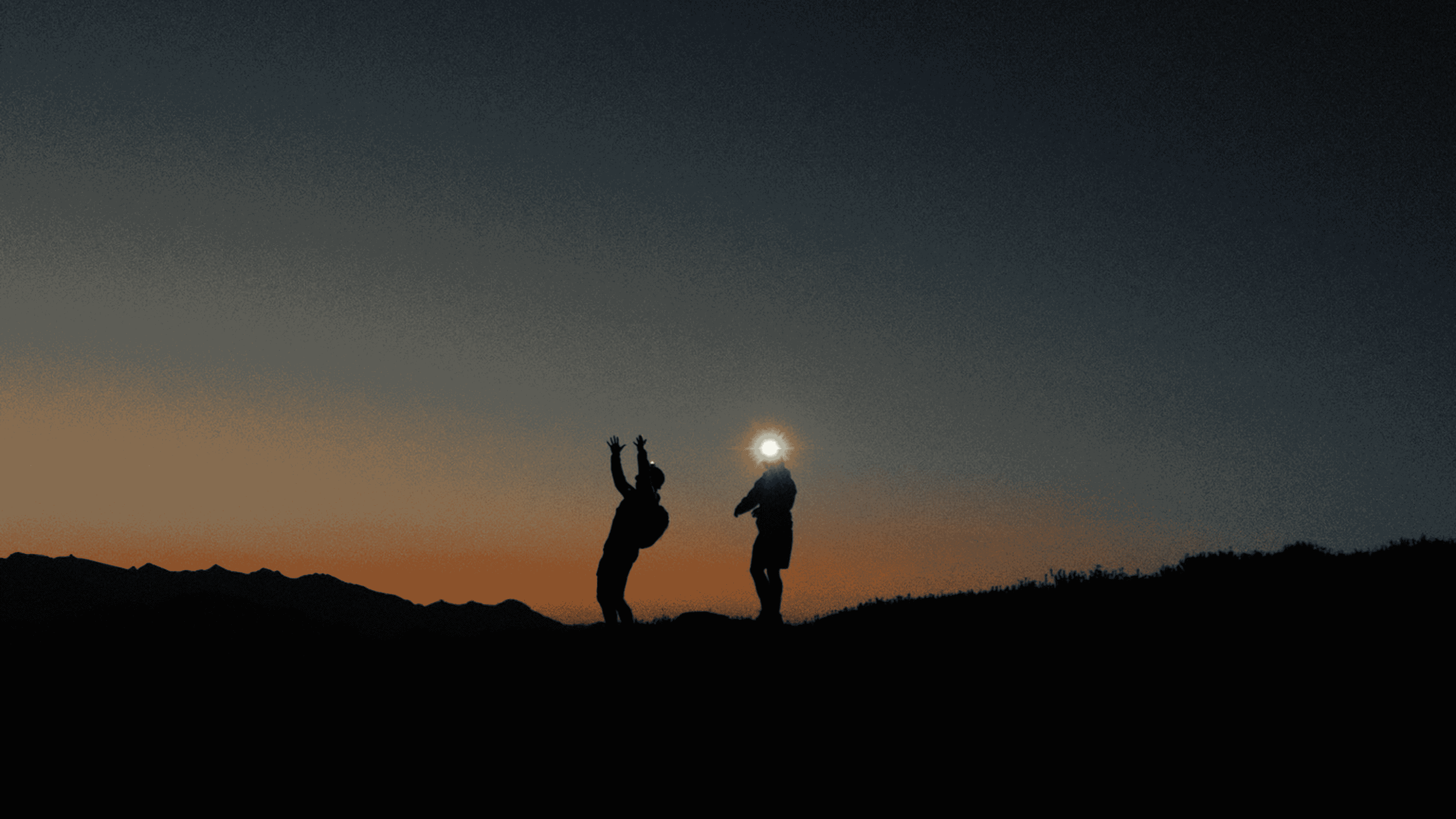 Silhouettes of two people standing on a hill during sunset, one person with arms raised and the other with a bright light behind their head.