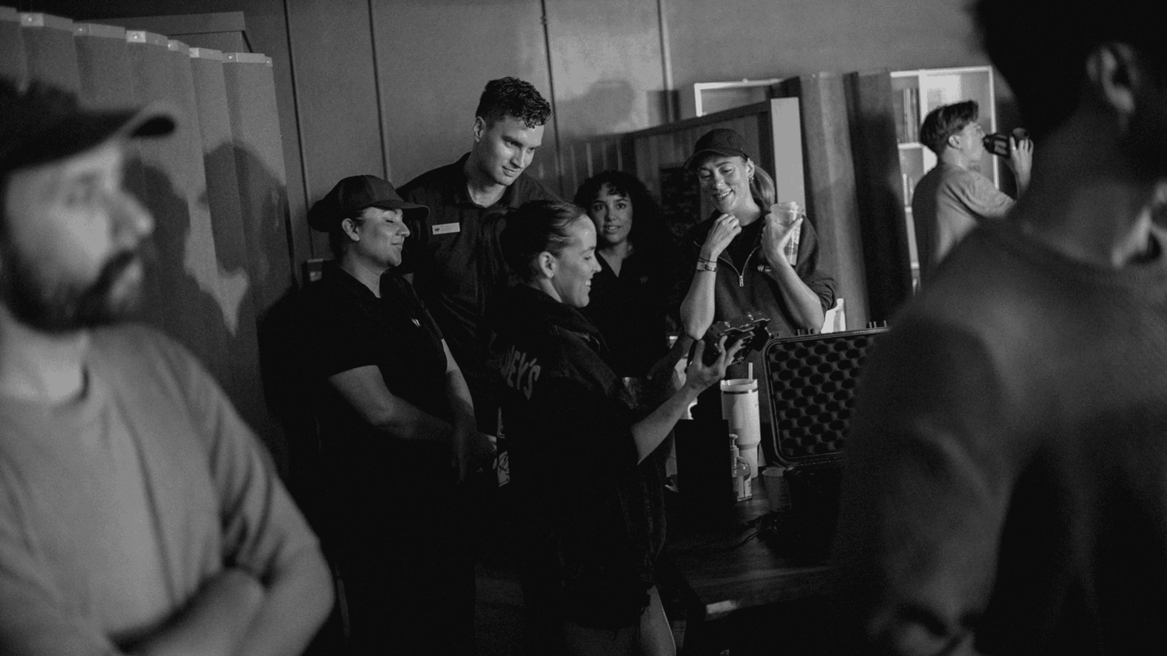 Group of people in a casual indoor setting, some looking at a device while others engage in conversation and one person drinks from a cup.