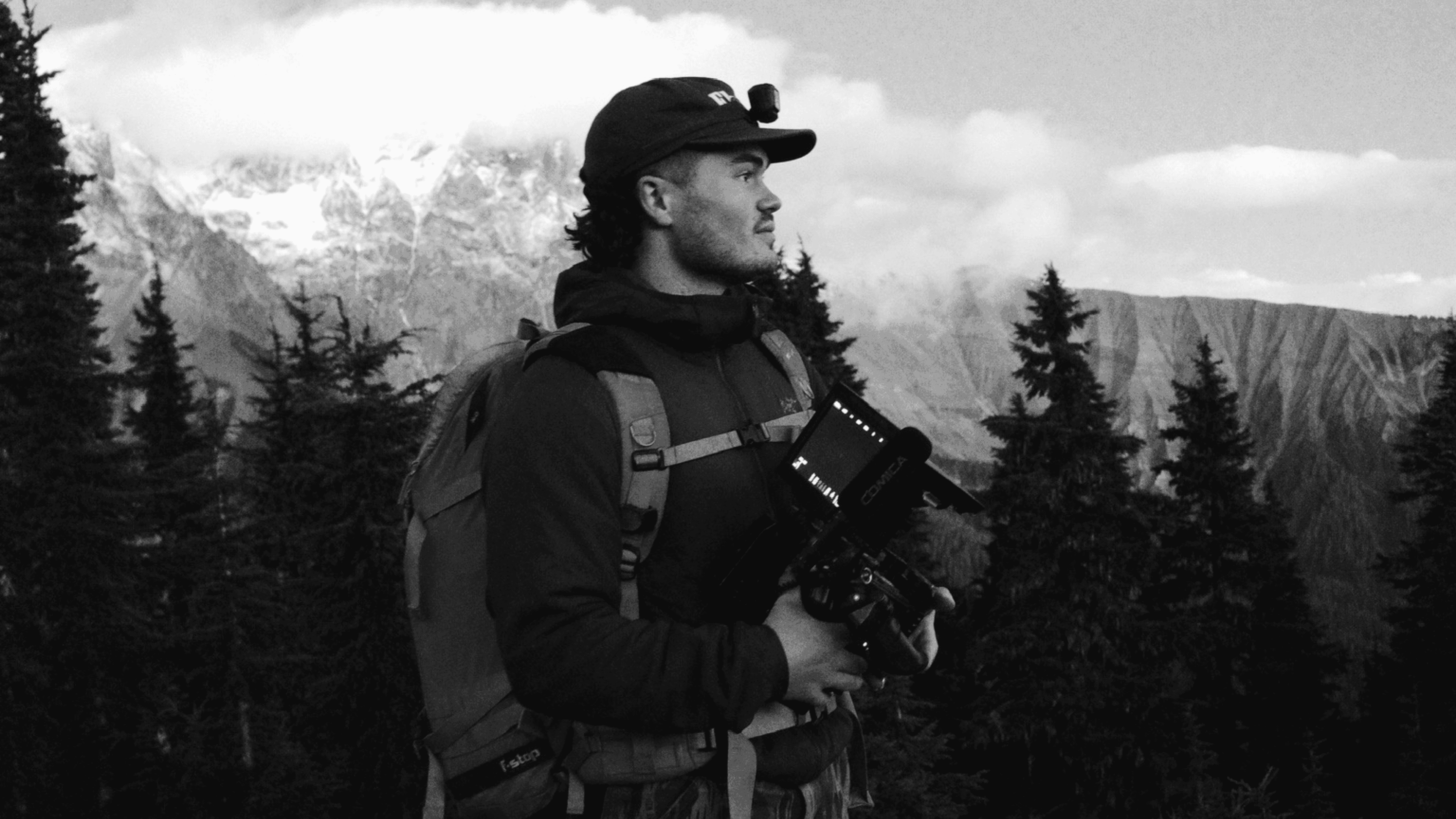 Man with a backpack and cap holding a professional camera against a mountainous forest backdrop.