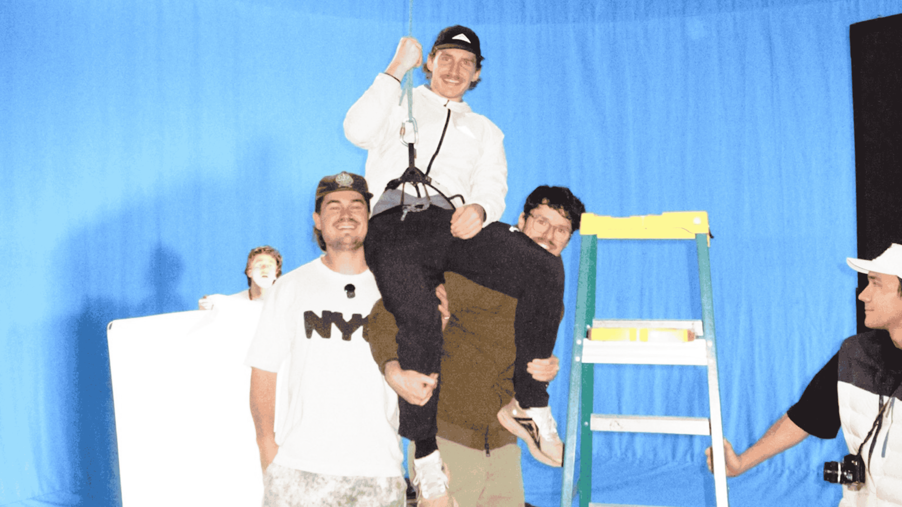 Four men posing in front of a blue backdrop; one man is being lifted by another, while one man holds a ladder and another stands nearby.