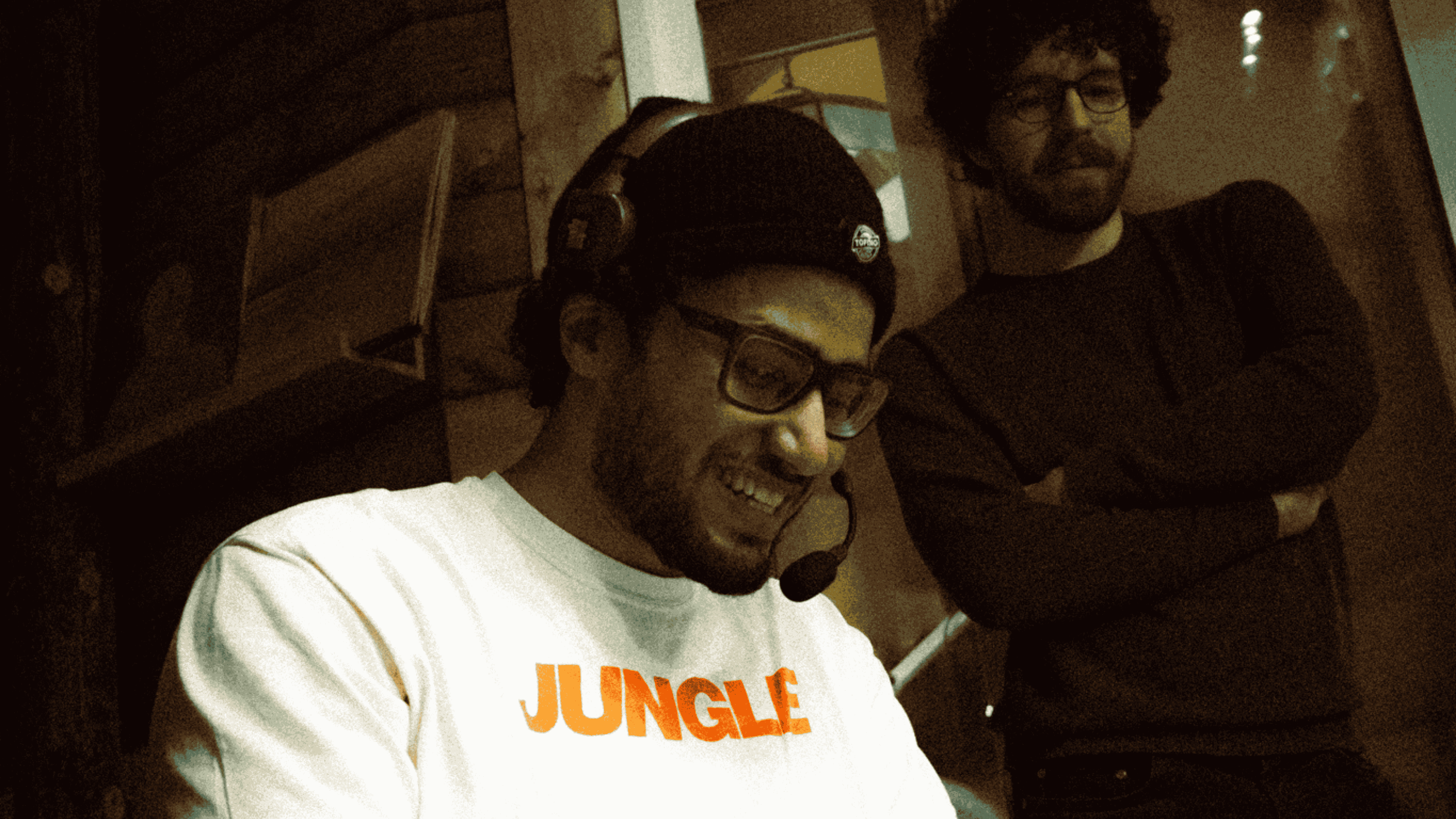 Two men indoors, one smiling wearing glasses, a black beanie, headset, and a white shirt with 'JUNGLE' in orange, the other man standing with arms crossed.
