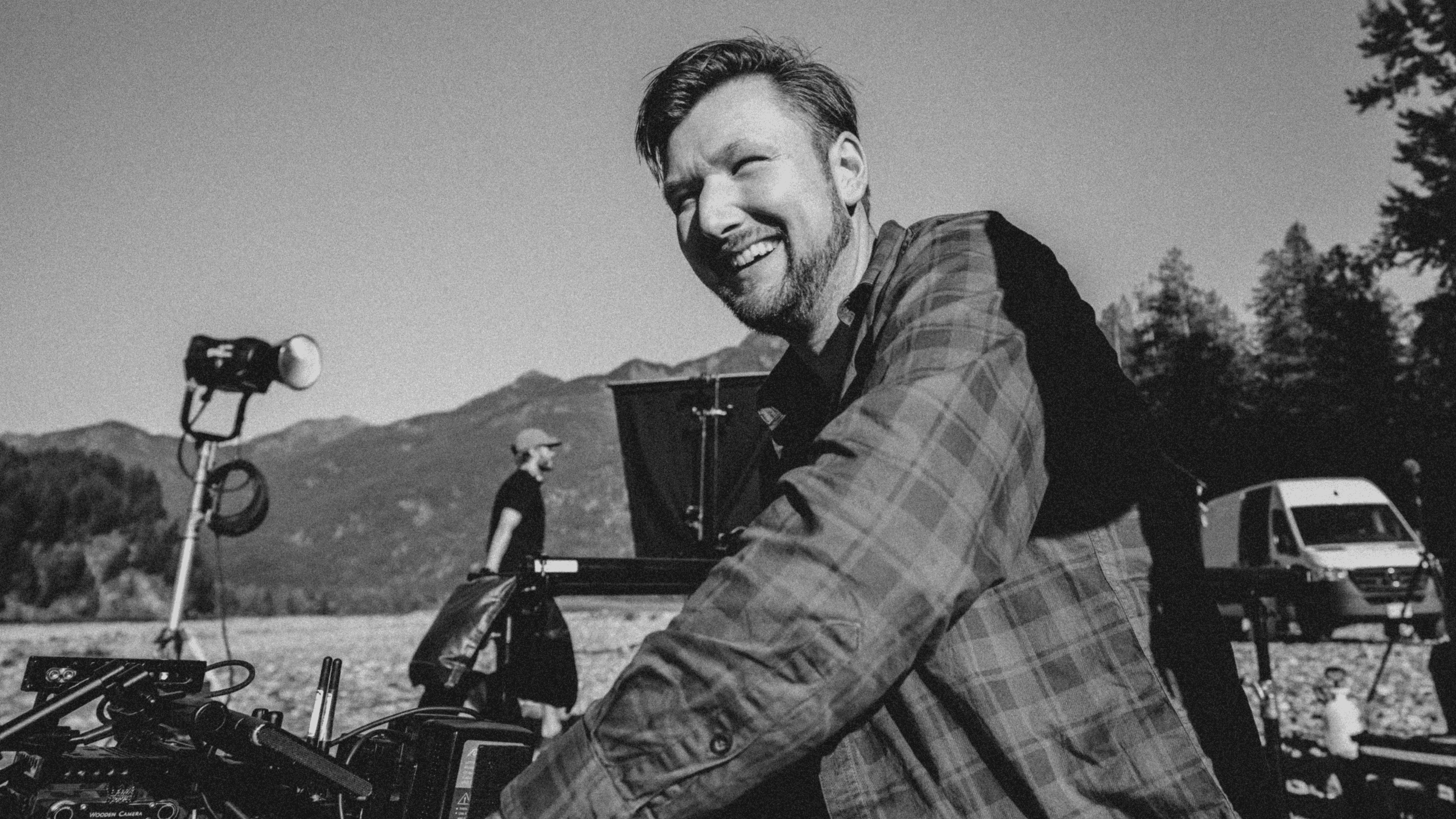 Smiling man in a plaid shirt operating a camera on a film set outdoors with mountains and crew in the background.